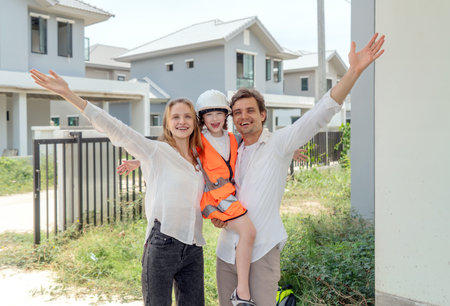 A cheerful family celebrate their new home in a developing neighborhood. The parent wear casual clothing while holding their child in a safety vest and hardhat, creating a lively atmosphere.の写真素材
