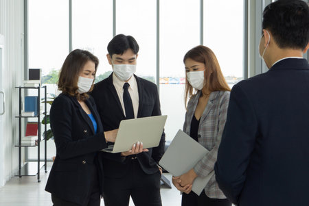 Four business professional in suit  collaborate on a project in a sleek office space. They are focused on a laptop screen while adhering to health protocol by wearing face mask.の写真素材