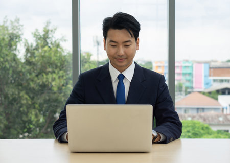 A professional man in a suit is seated at a large desk, intently working on a laptop.の写真素材