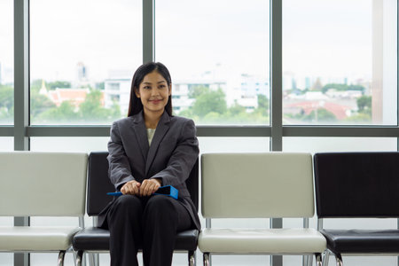 Young asian businesswoman in gray suit sit on a bench near the window, There is a blue file on her...