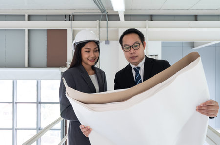 Asian businessman in black suit holding construction drawing. Young female engineer stand next to...