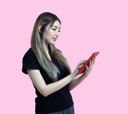 Young asian woman in casual clothing, holding and looking at a tablet computer. Portrait on a simple pink background with studio light. Isolatedの写真素材