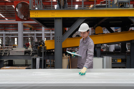 Young asian worker in safety helmet and protective gloves checking material covered in plastic. Work atmosphere in metal sheet manufactory.の写真素材