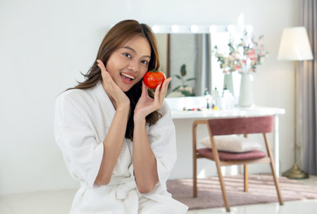 Young asian woman dressed in comfortable robe holding tomato, smile while sitting in front of vanity mirror table at dressing room.の写真素材