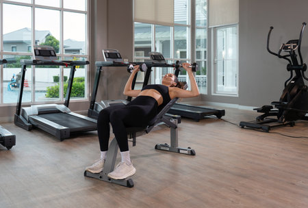 Young woman in sport clothing lean on the bench in a contemporary gym, perform a incline dumbbell press. Natural light stream through large window, illuminating the fitness space.の写真素材