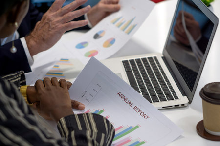 Team member are reviewing an annual report, discussing data and graph. Colorful chart are spread on the table alongside a laptop and a coffee cup.の写真素材