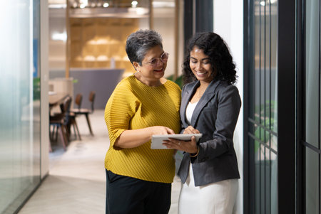 In a contemporary office, two women engage in a friendly discussion while examining a tablet together. The atmosphere is vibrant and professional, showcasing teamwork.の写真素材