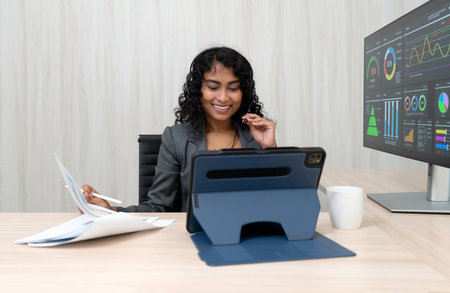 A businesswoman with curly hair smile as she examine data on her tablet while sitting at a desk in a sleek office environment. She has document and a coffee mug nearby.の写真素材