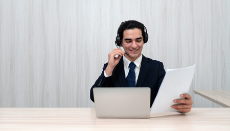 A young man dressed in a suit participate in a virtual meeting, wearing headphone and smiling as he...