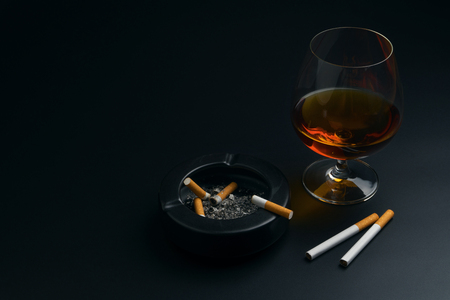 a glass of brandy, cigarettes and black ceramic ashtray full of ashes with the cigarette butt, on the black tableの写真素材
