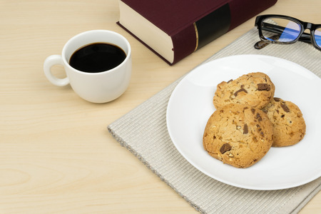 a cup of coffee, three pieces of chocolate chip cookies in a white round dish, a red book and an eyeglasses on the wooden table.の写真素材