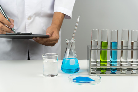 blue liquid flask with glass stirring rod, test tubes in the rack, watch glass and beaker on the table and a scientist writing test results in the laboratoryの写真素材