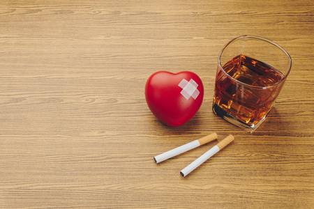 unhealthy lifestyle concept, red heart symbol with adhesive plaster, two cigarettes and a glass of bourbon whiskey on wooden tableの写真素材