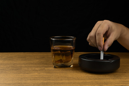 hand stubbing out a cigarette on the black ceramic ashtray, a glass of bourbon whiskey on wooden table.の写真素材