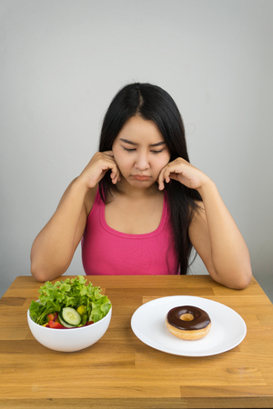 beautiful plus size Asian woman choosing between salad and chocolate doughnut, dieting conceptの写真素材