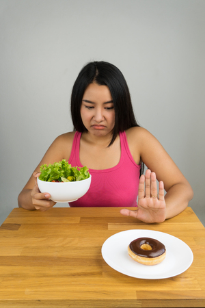 beautiful young Asian woman choosing salad bowl and rejecting chocolate doughnut, dieting conceptの写真素材
