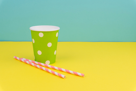 green polka dot party paper cup and pink striped straws on yellow table with blue backgroundの写真素材