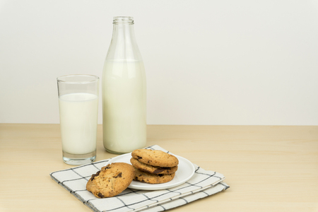 a glass of milk with milk bottle and the chocolate chip cookies in a white round dish on the wooden table.の写真素材