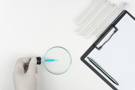 laboratory scene, the scientist holding a blue liquid dropper over the Petri dish, test tubes and a clipboard with pen on the white table.の写真素材