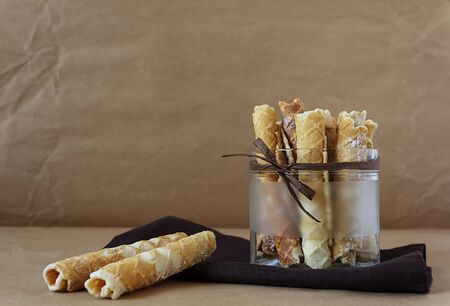Homemade baking. Delicious wafer rolls with sugar in a glass bowl with decorative decoration on a dark brown textile linen napkin on a craft paper background. Sweet, baked tubes. Food photo, copy spaceの写真素材