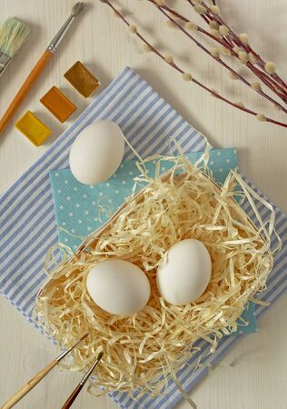 Preparations for the celebration of Easter, spring mood. Whole raw white eggs prepared for coloring and decorating on straw with brushes, watercolor paints and budding willow twigs . Cozy home pleasant pastimeの写真素材