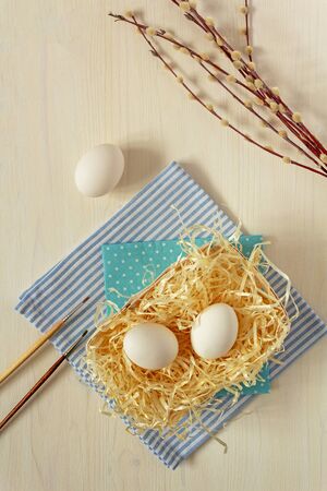 Fresh, raw, white eggs on straw with brushes, watercolor paints and willow twigs on a wooden background with copy spaseの写真素材