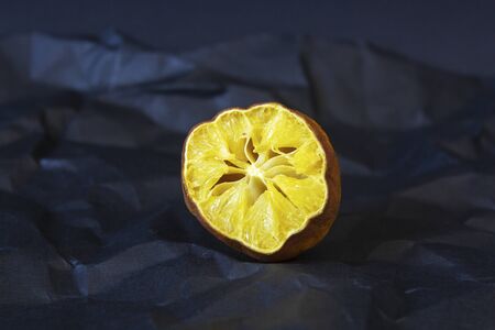 Ugly organic lemon. Deformed half of a lemon with old flesh with damaged, rotten ugly peel on a black crumpled paper background with large creases. Horizontal orientation Organic wasteの写真素材