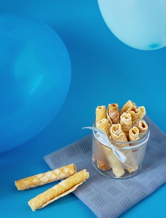 Children's holiday at home. Homemade cakes, sweet waffle tubes with powdered sugar in a glass bowl with balloons on a blue background. Food photos, vertical orientationの写真素材