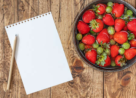 A bowl of ripe useful gooseberries, strawberries on wooden background with a sheet of white paper and a pencilの写真素材