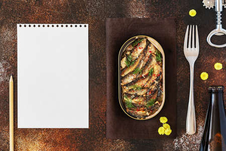 Open tin of Baltic sprats in oil with spices with a fork and a bottle of dark beer with a sheet of white writing paperの写真素材