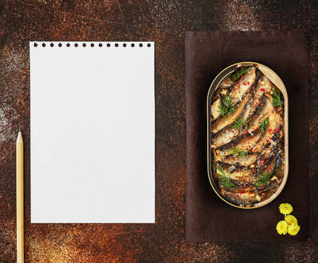 Open tin of Baltic sprats in oil and spices with a sheet of white writing paper, pencil and yellow flowers on an old brown background. Place to record a fish snack recipe. Food photo, top viewの写真素材