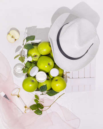 Cosmetic composition with green apples in a wooden box, a summer hat and bottles with cream on a white background. Healthy, summer, natural cosmetics. Anti-aging treatments for summer seasonの写真素材