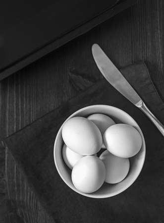The concept of a simple healthy natural food. Chicken, white, fresh farm eggs in a white bowl prepared to cook an omelet on a dark wooden background. Black and white food photo, vertical orientationの写真素材