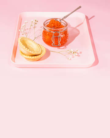 Vertical romantic food composition with a jar of salmon red caviar, tartlets and flowers on a tray on a pink background with copy space. Appetizer with sea delicacies. Gourmet sea foodの写真素材