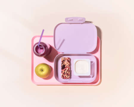 The concept of healthy school meals. Food composition with lunch box with yogurt, cereal bar, apple and oatmeal berries cocktail on a pink background with window light. Flat lay, horizontal bannerの写真素材