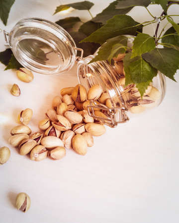 Vertical, natural, nut, food composition with a glass jar with roasted, salty pistachios and leaves on a white background with copy space. Healthy, tasty snacks. Summer beer party in the open airの写真素材