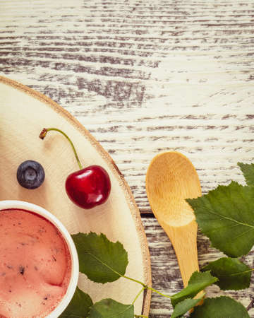 Summer, food composition with natural, vegan ice cream, blueberries, cherry, leaves and wooden spoon on white wooden background. Eco friendly still life with berry, frozen yogurt. Healthy, tasty snackの写真素材