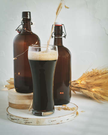 Vertical, food composition with glass of cold, dark, unfiltered beer, two dark brown bottles and bouquet of wheat on a white background. The concept of natural beer made in small home breweriesの写真素材