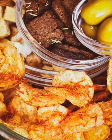 Rye crackers, wheat crackers, potato chips with spices and green olives in glass bowls on a white tableの写真素材