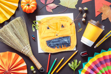 Autumn flat lay with children drawing of a halloween pumpkin, color pencils and autumn decorations on a black chalkboardの写真素材