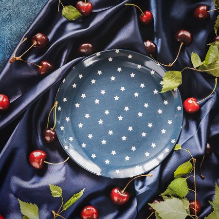 Dark blue composition with an empty blue plate with a star pattern and cherry berries with leaves on a satin fold fabric on a blue background. Creative still life with empty plate. Flat layの写真素材