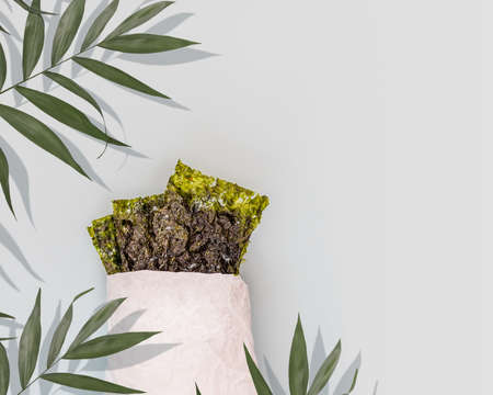 Salty dried seaweed snack in a white paper bag with palm leaves on a blue background with copy space. Healthy sea snack suitable for keto and vegan diet. Japanese food, chips of nori seaweedの写真素材