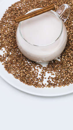 Non lactose buckwheat milk. Vegan food composition with glass cup of plant milk with a cinnamon stick on a white plate with buckwheat groats on a white table. Vegan healthy milk productの写真素材