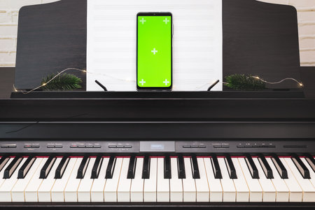 Smartphone with green screen on a electronic piano with winter holiday decoration. Piano lesson online. Online music holiday concert or lecture. Mockup for mobile music apps. Online music educationの写真素材