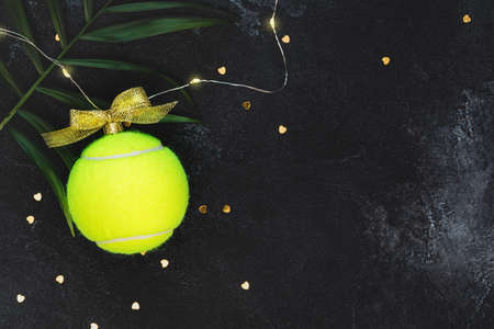 Tennis, winter holidays composition with yellow tennis ball in the form of a New Year's ball, palm leaves and confetti on a black background with copy space. New year banner for a tennis competitionの写真素材