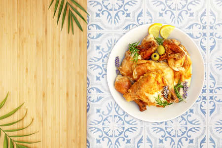 Homemade grilled chicken with crust, sprigs of rosemary, lavender flowers, lemon slices and olives on a ceramic tile table and wooden kitchen board with palm leaves and copy space, flat layの写真素材