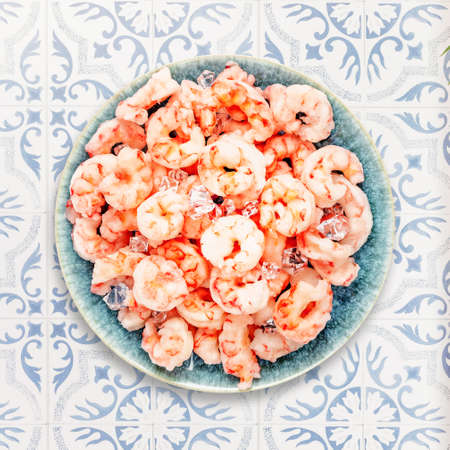 Close up natural frozen pilled shrimps with ice cubes on a blue plate on a tile ornamental table. Prawns cooking. Frozen foods. Mediterranean, keto dietの写真素材