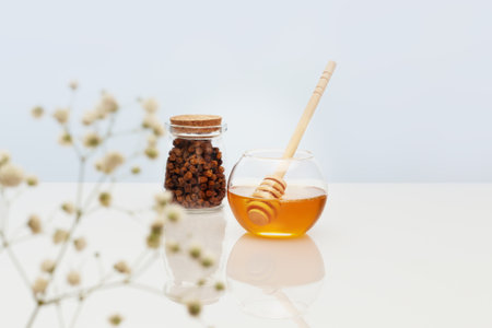 Healthcare composition with propolis and honey in glass jars with honey dipper on a white table with reflection and flowers. Bee products are an natural, folk medicine. Immune boosting productsの写真素材