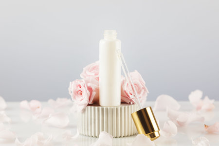 White dropper bottle with serum on a marble podium on a white table with tender pink roses and rose...