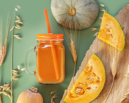 Eco friendly card with glass of pumpkin juice with straw, pumpkins, rye and wheat ears on a green background. The new crop of pumpkins. Autumn harvest of wheat, rye and pumpkins on a table. smoothiesの写真素材